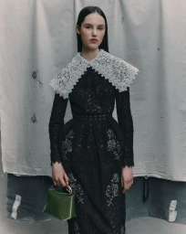 Model wearing a black lace dress with an ornate white collar and holding a green handbag against a textured backdrop