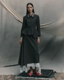 Model standing in a dark tailored peplum suit with embroidered collar against a grey draped backdrop