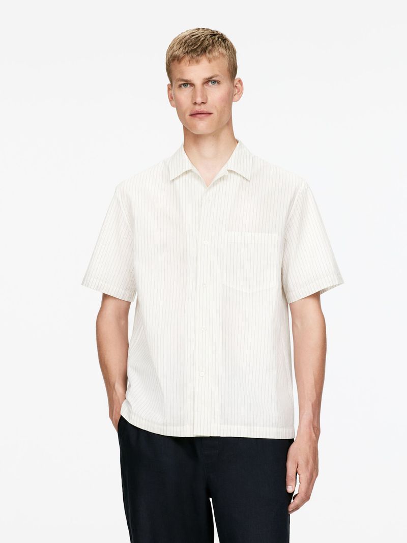 Man in a striped short-sleeved shirt and dark pants poses in front of a plain white background. Fashion portrait. Studio lighting.
