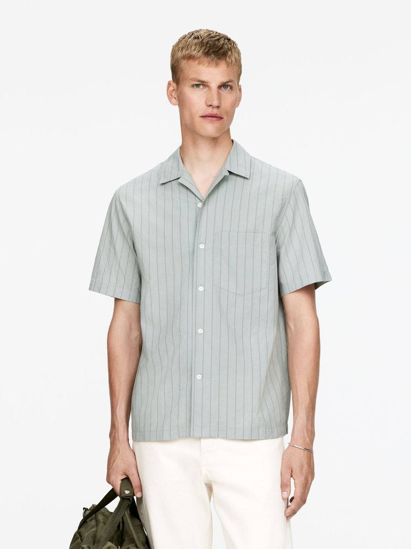 Adult male in a light green, striped short sleeve shirt and cream pants, standing indoors while holding a dark green tote bag.