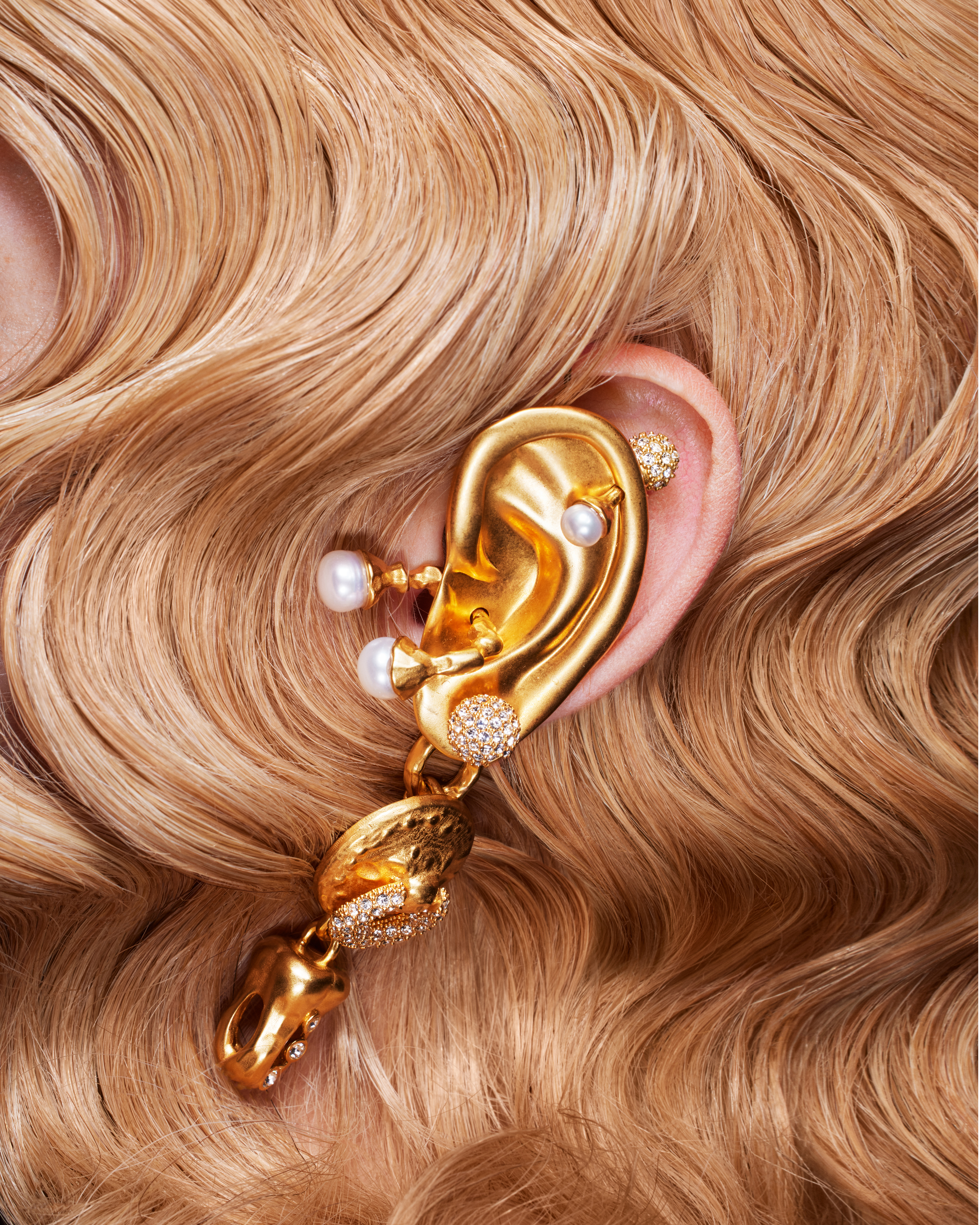 Close-up of a human ear adorned with an elaborate gold-toned metal earring featuring pearls, rhinestones, and pendant charms.