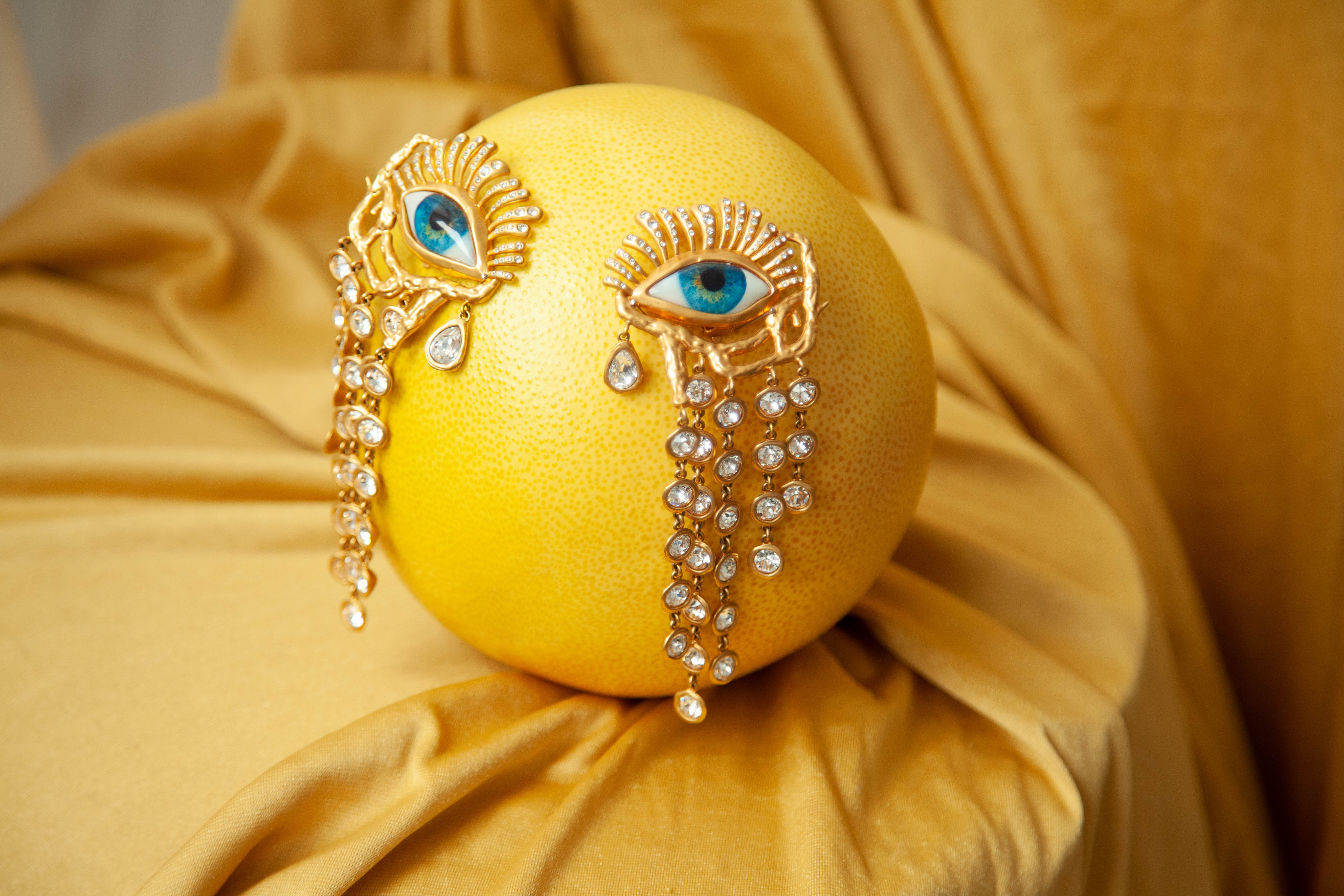 Still life of a pomelo with two golden 'eye' brooches adorned with clear gems and blue glass pupils, set on golden satin fabric.