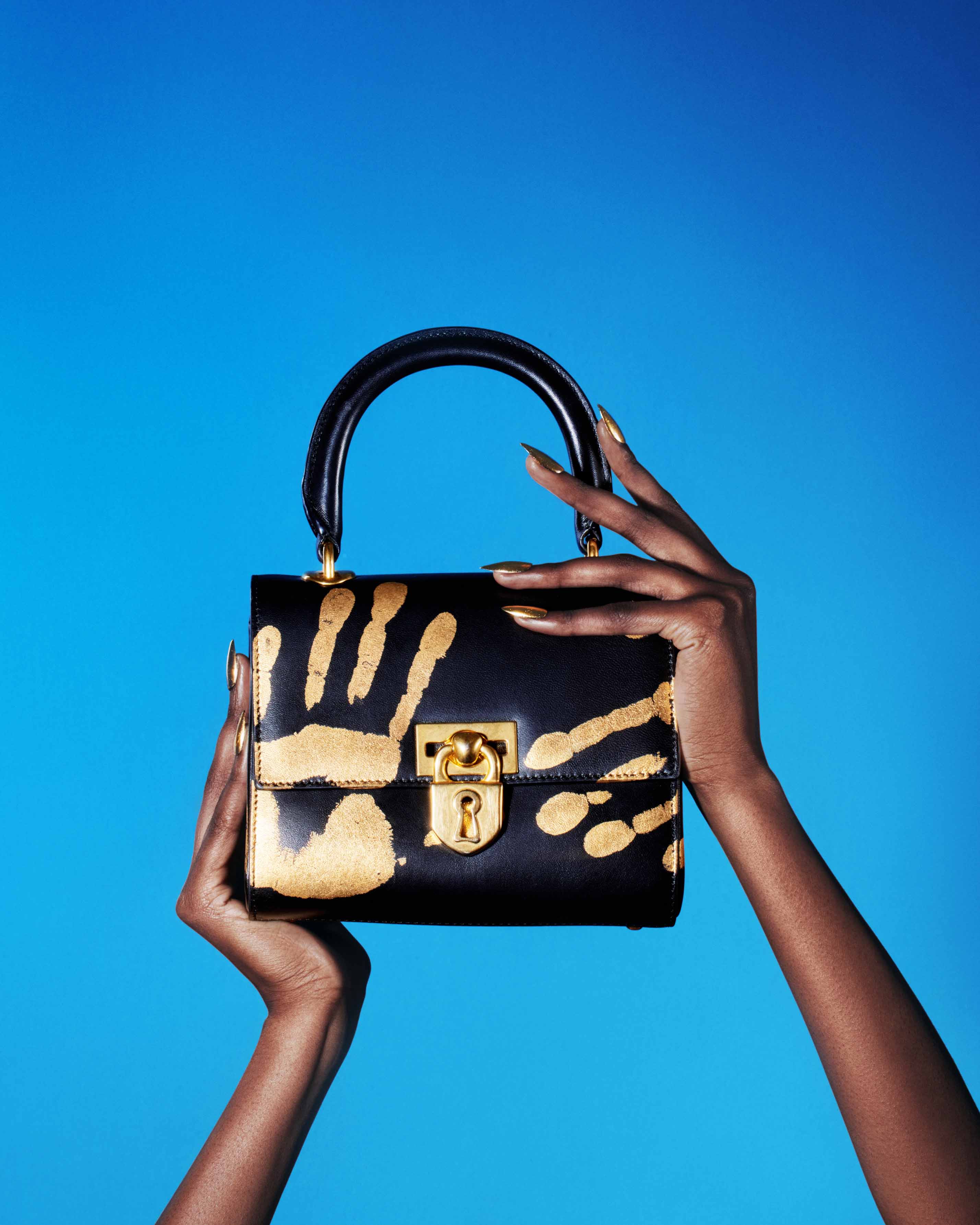 A black leather handbag with gold handprint details is held up by a woman's hands against a bright blue background in a studio shot.