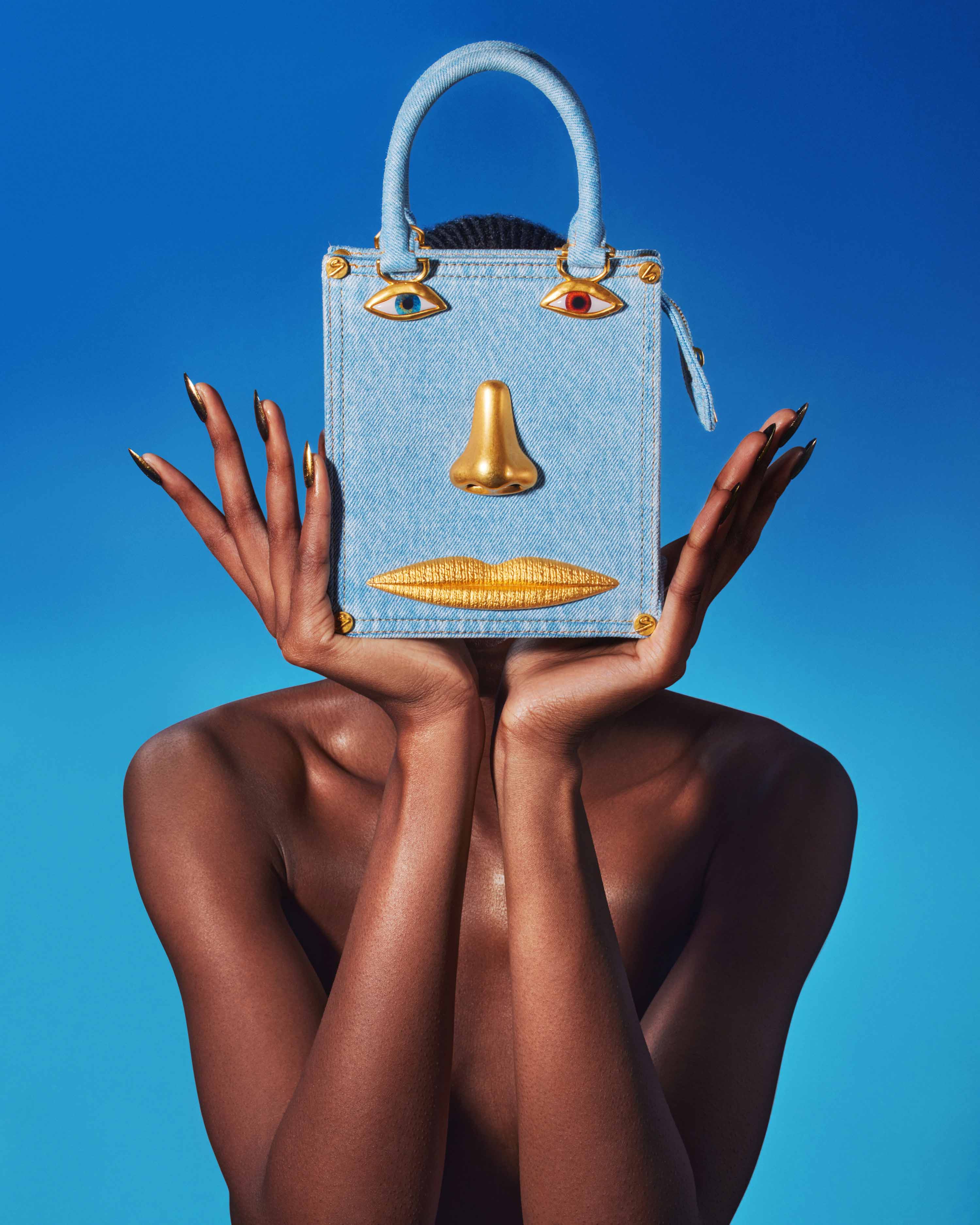 Denim purse with a face design held by a dark-skinned woman with gold fingernails set against a clear blue sky backdrop.
