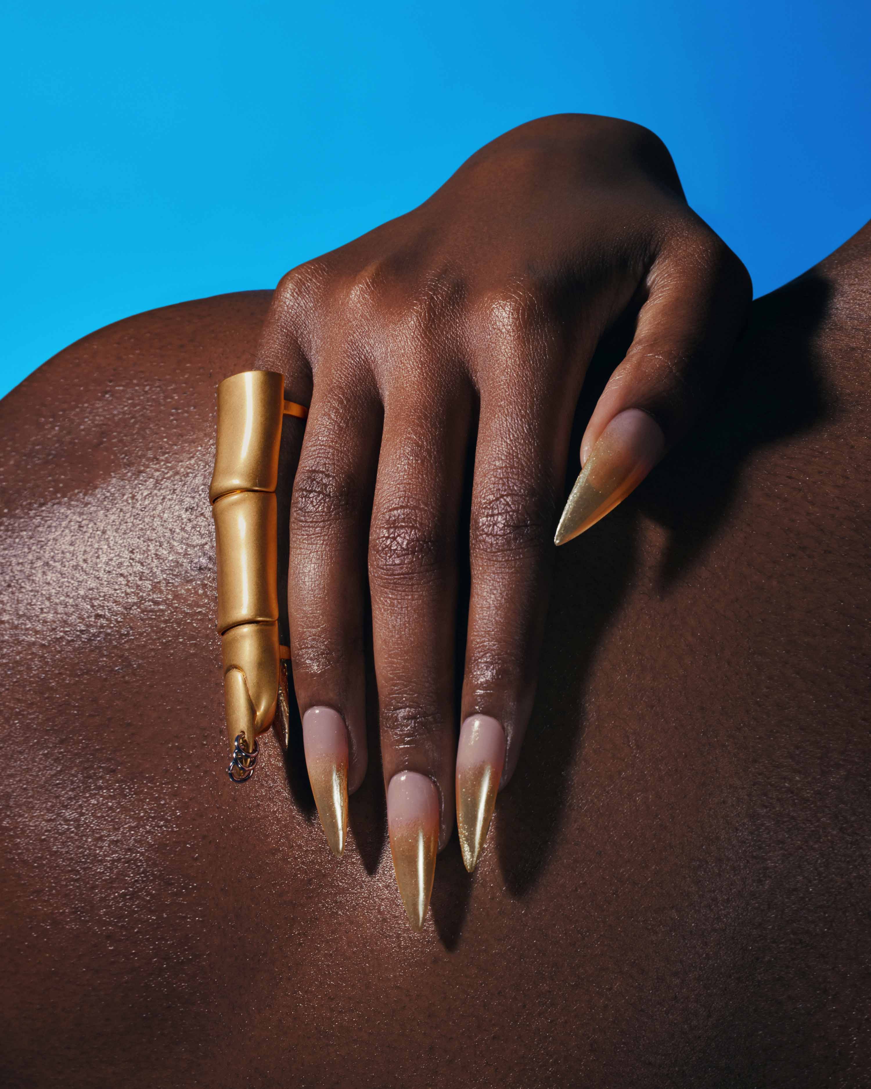 Dark-skinned arm and hand showing long, pointed, gold-tipped nails and bamboo-shaped ring, against a vivid aqua blue background.
