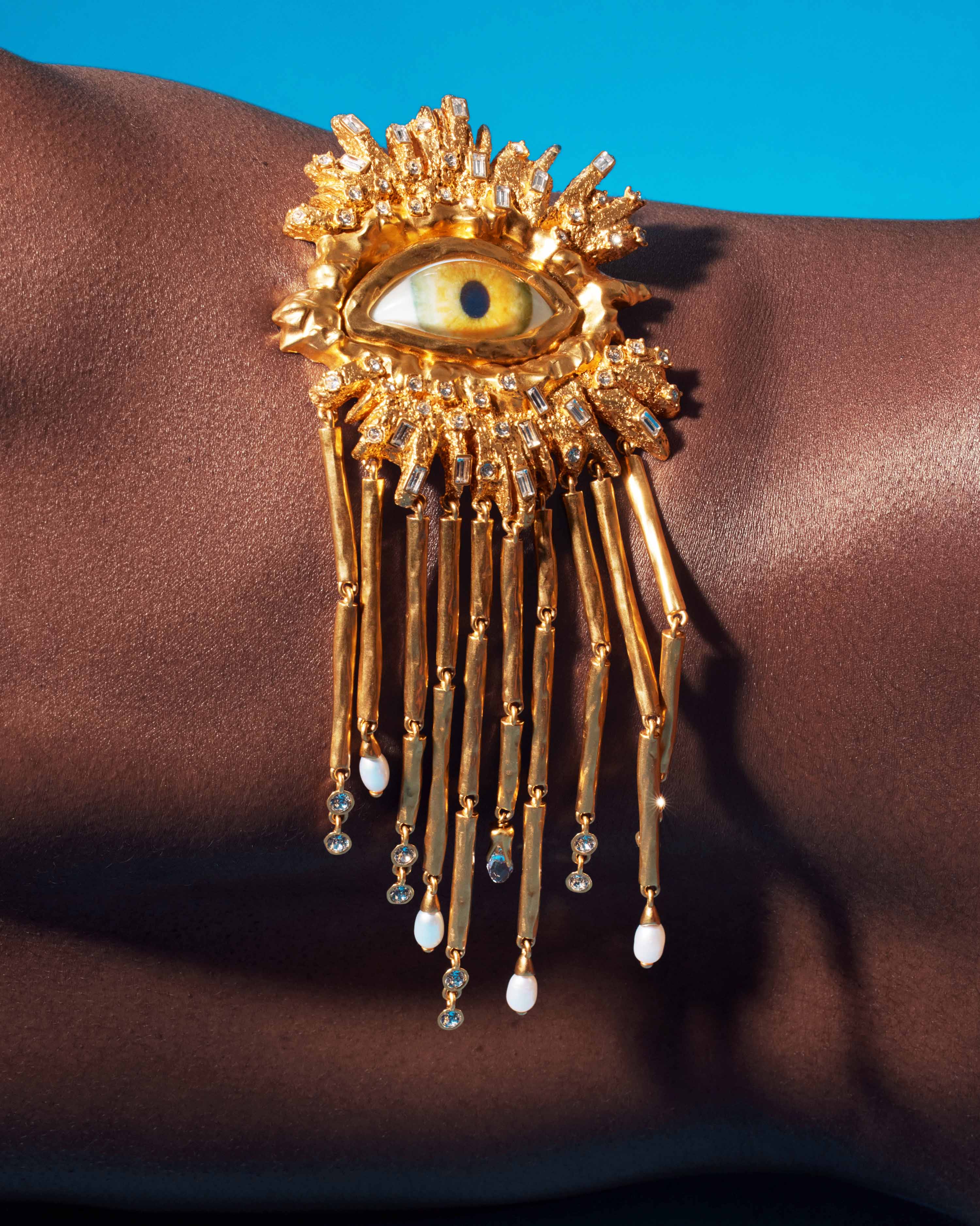 Close-up of dark-skinned person wearing a large gold eye brooch with gold rods, crystals, and pearls on the right shoulder against a blue background.