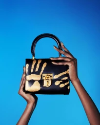 Dark-skinned hands holding a black leather handbag with a gold handprint motif against a bright blue background