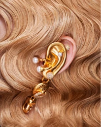 Close-up portrait of an ear adorned with an oversized sculptural gold ear cuff featuring pearl accents and dangling elements, set against wavy blonde hair