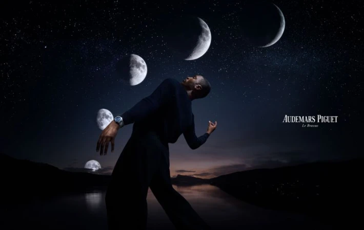 Man in dark clothing gazes upwards with multiple moons in a night sky, watch visible on wrist, against a dark, reflective landscape.