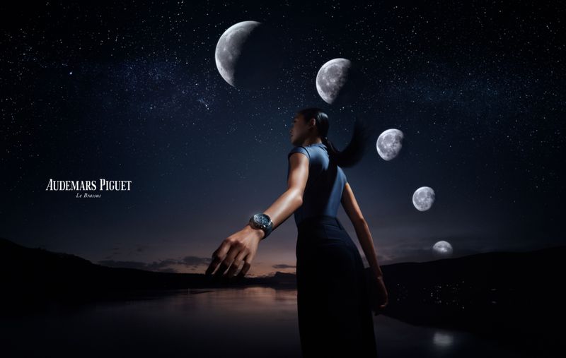 Woman stands on a shoreline wearing a watch, with moon phases and stars overhead against a mountainous horizon in a watch advertisement.