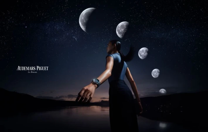 Woman stands on a shoreline wearing a watch, with moon phases and stars overhead against a mountainous horizon in a watch advertisement.