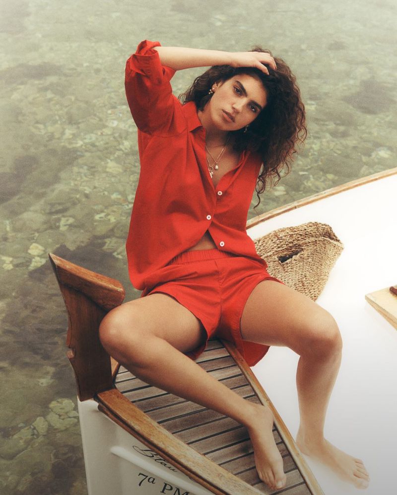 Brunette woman poses in a red linen shirt and shorts on a boat, with clear water visible in the background, in a medium shot.