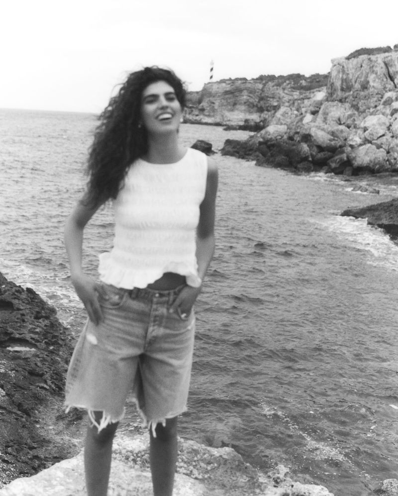 Monochrome shot of smiling woman with curly hair in top and denim shorts, standing on rocks with ocean and coastline in the background.