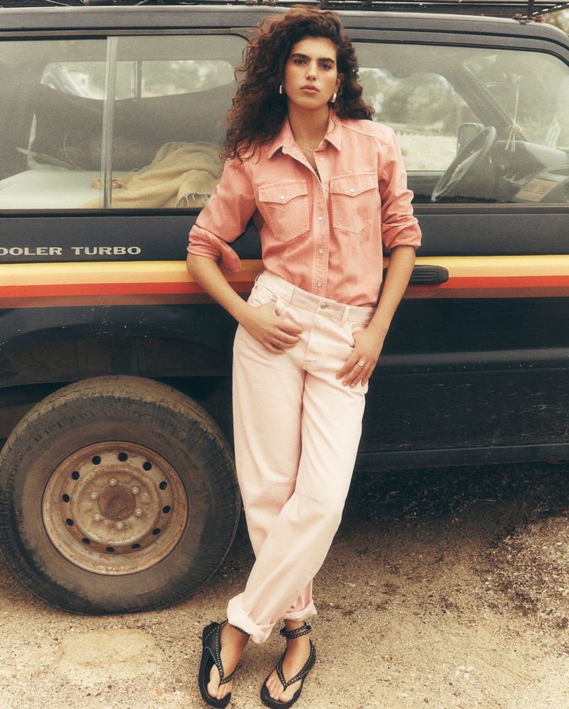 Woman in pink shirt and pants poses next to a black SUV with an orange stripe. She is wearing flip-flops, and the background is neutral.