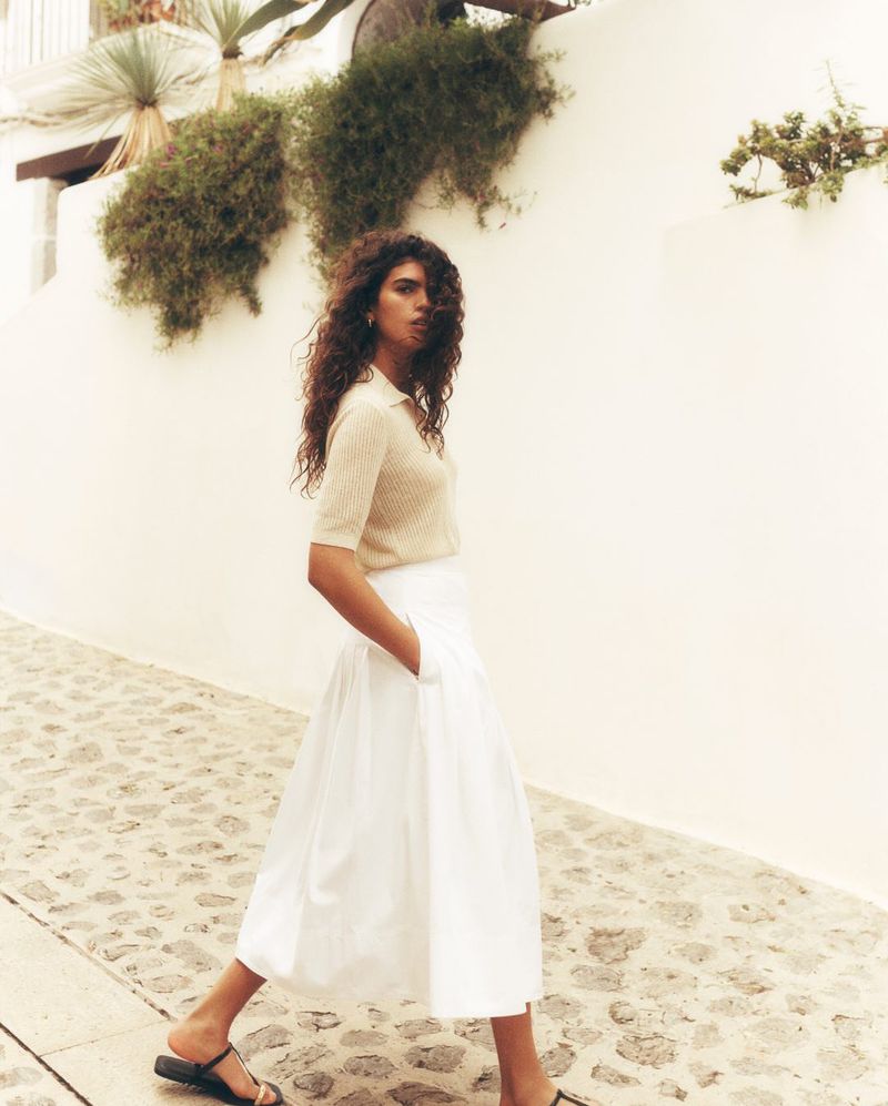 Woman in a skirt and top walking along a stone path. The background features a white wall and greenery against a bright sky.