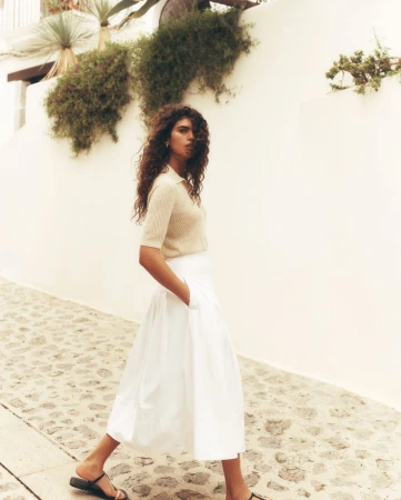 Woman in a skirt and top walking along a stone path. The background features a white wall and greenery against a bright sky.