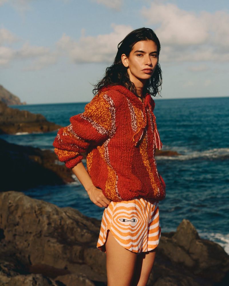 Adult woman poses in a knitted sweater and shorts on a rocky beach with the sea and cloudy sky in the background on a sunny day.