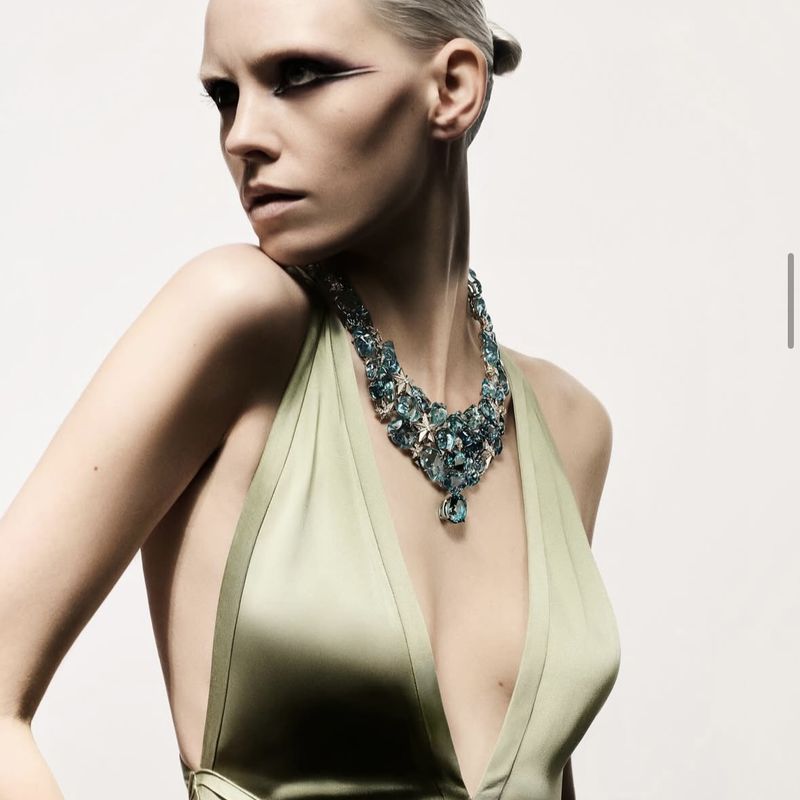 Elegant woman with smoky eye makeup poses wearing a green satin dress and a statement teal gemstone necklace on a white backdrop.