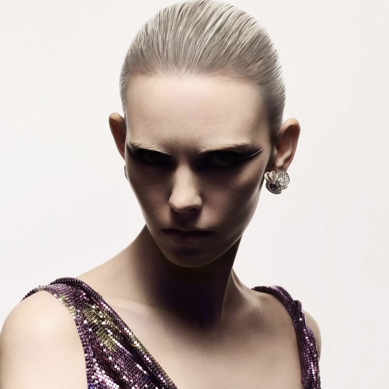 Close-up portrait of a striking woman with slicked-back hair and statement eye makeup wearing a sparkling purple dress and statement earrings.