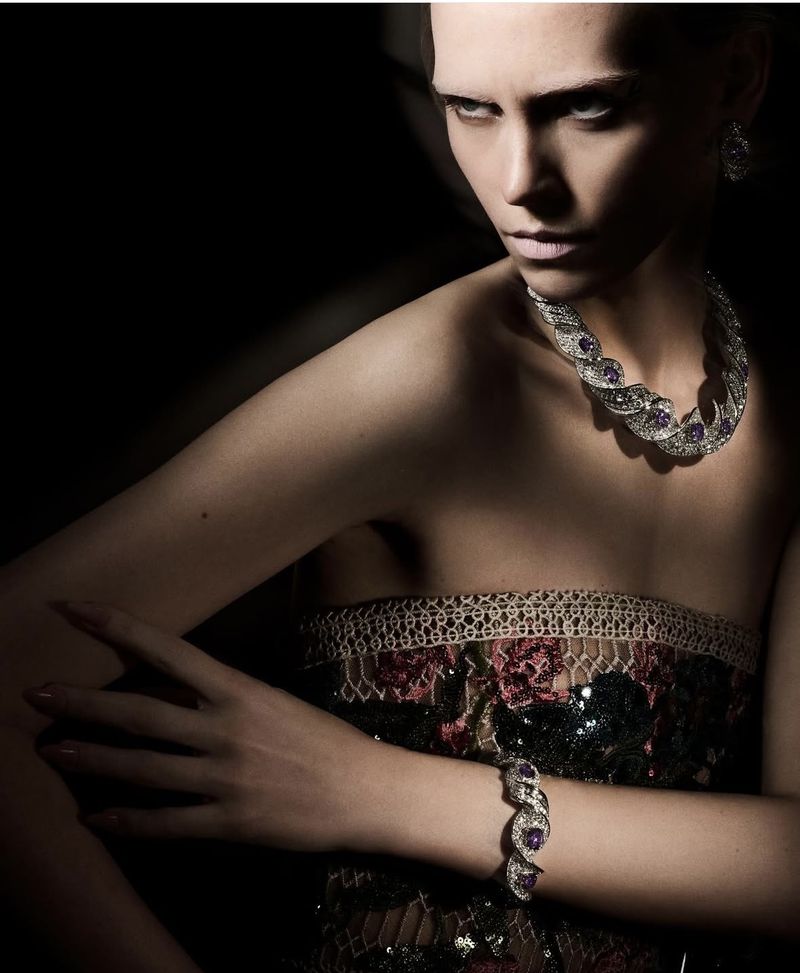 Woman wearing a floral dress and jewelry including a necklace and a bracelet posing in front of a dark background.