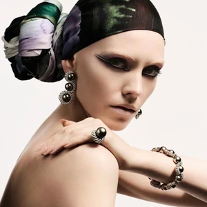 Model wearing a printed headscarf, dark eye makeup, and pearl jewelry against a plain background