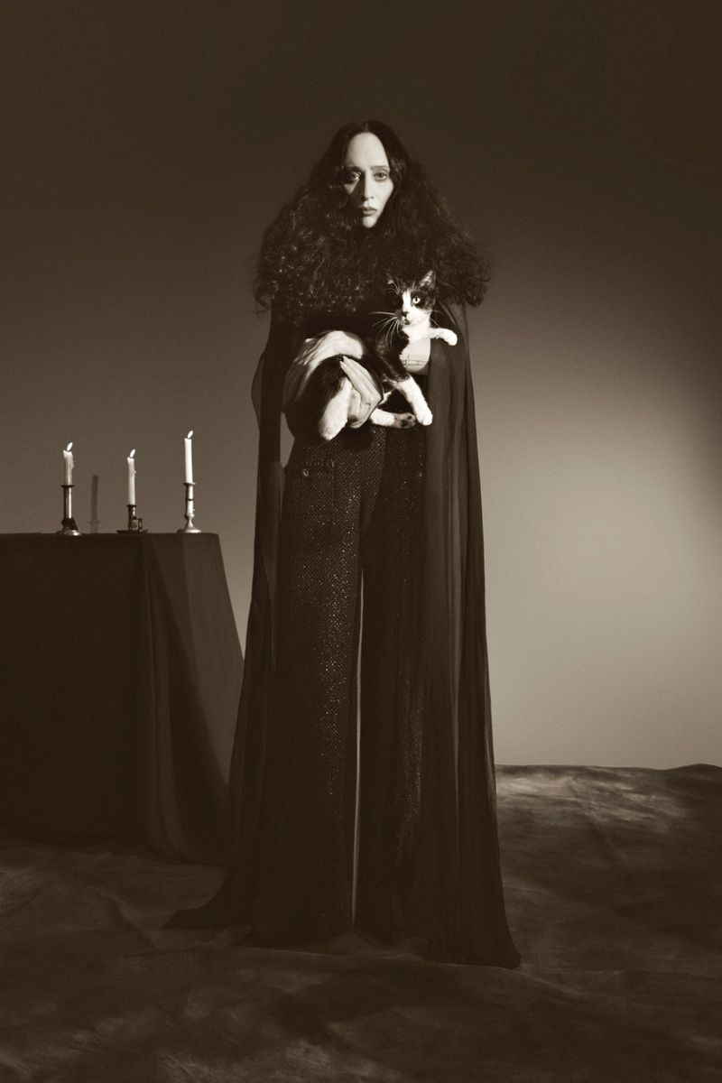 A woman in a dark outfit holds a cat, standing in front of a candlelit table, casting a sepia-toned, shadowy and mysterious ambiance.
