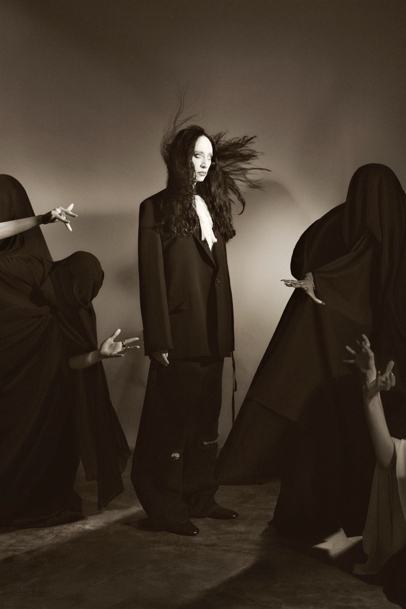A single adult female in a blazer and long skirt stands in the center, surrounded by four figures cloaked in dark fabric in a photo studio.