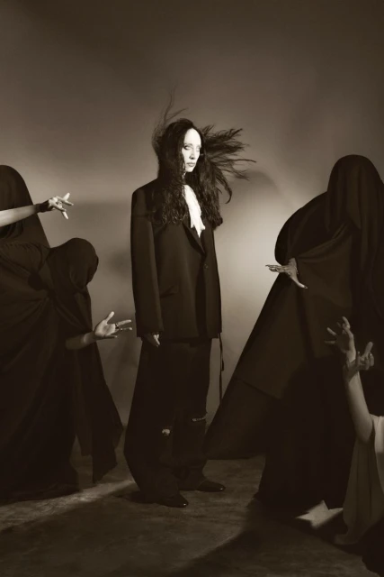 A single adult female in a blazer and long skirt stands in the center, surrounded by four figures cloaked in dark fabric in a photo studio.