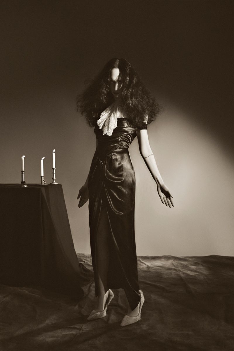 A woman with dark hair and a long black dress stands near candles in a dimly lit studio, creating a dramatic, sepia-toned fashion shot.