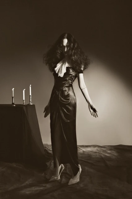 A woman with dark hair and a long black dress stands near candles in a dimly lit studio, creating a dramatic, sepia-toned fashion shot.