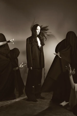 Model Talytha Pugliesi stands centrally flanked by figures in dark robes in a monochromatic editorial fashion photograph.