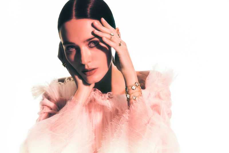 Close-up portrait of an adult woman with a light skin tone in a ruffled pink top wearing rings and bracelets, against a white backdrop.