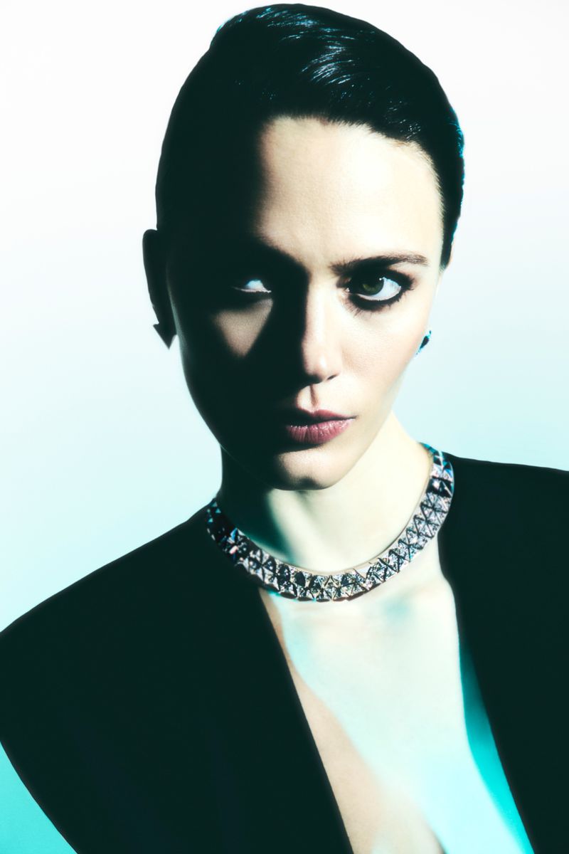 Close-up portrait of a woman with dark hair wearing a sparkling necklace and a blazer, set against a cool white and blue background.