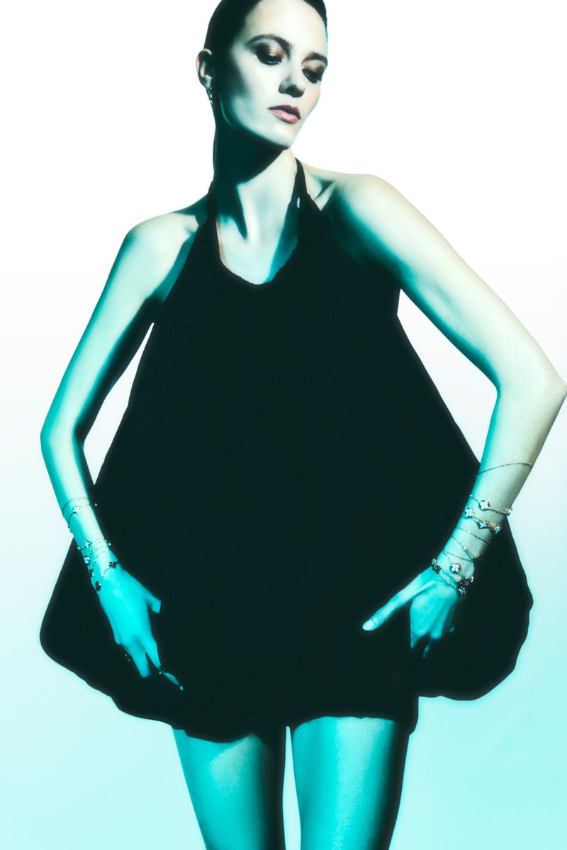 Fashion model poses in a short black halter dress with arms outstretched, wearing bracelets, against a blue-white gradient background.