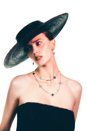 Model wearing a wide-brimmed black hat and layered pendant necklaces against a plain white background