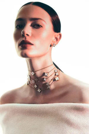 Close-up portrait of a woman wearing layered gold necklaces and matching stud earrings against a bright white background.