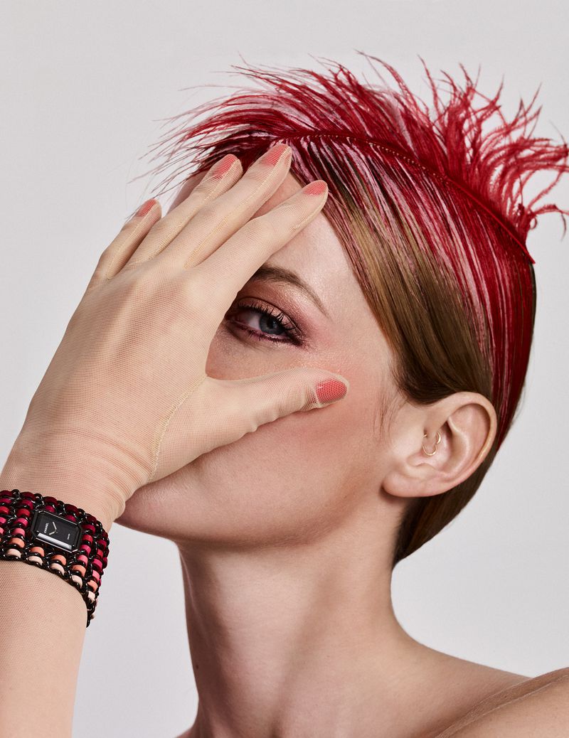 Close-up portrait of a fair-skinned woman with red feathered hair, wearing a bracelet watch and glove, hand partially obscures face.