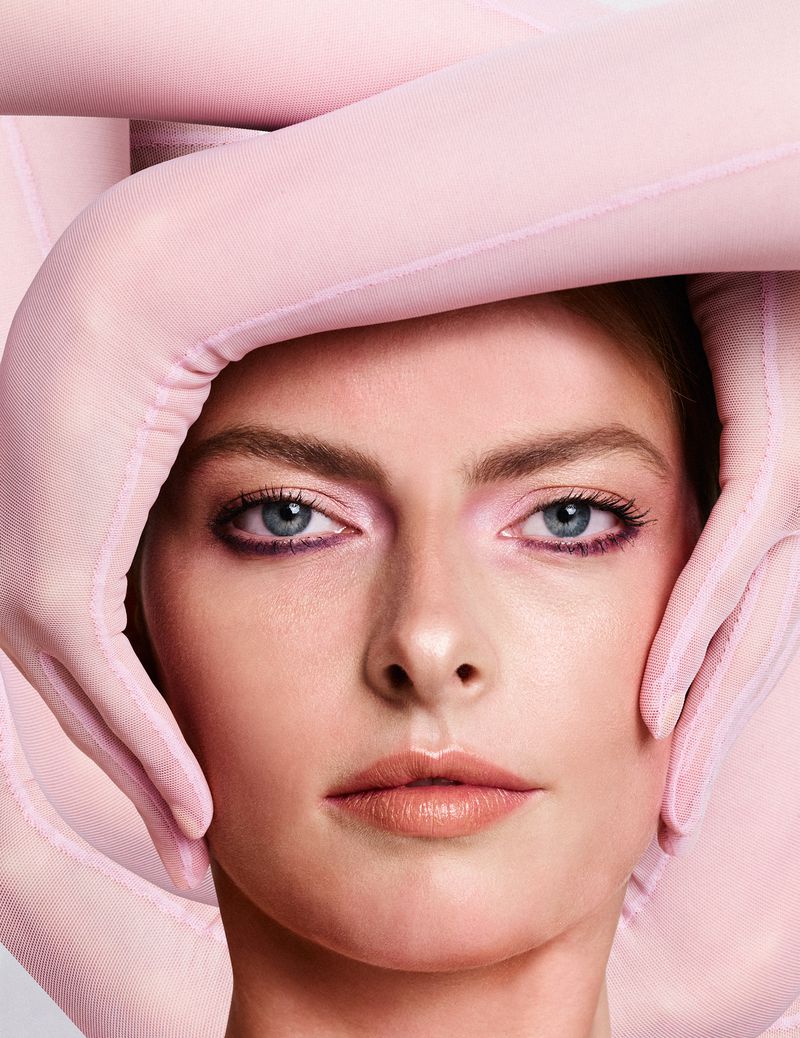 Close-up portrait of a young woman with blue eyes and light makeup, hands covered in pink semi-sheer fabric framing her face.