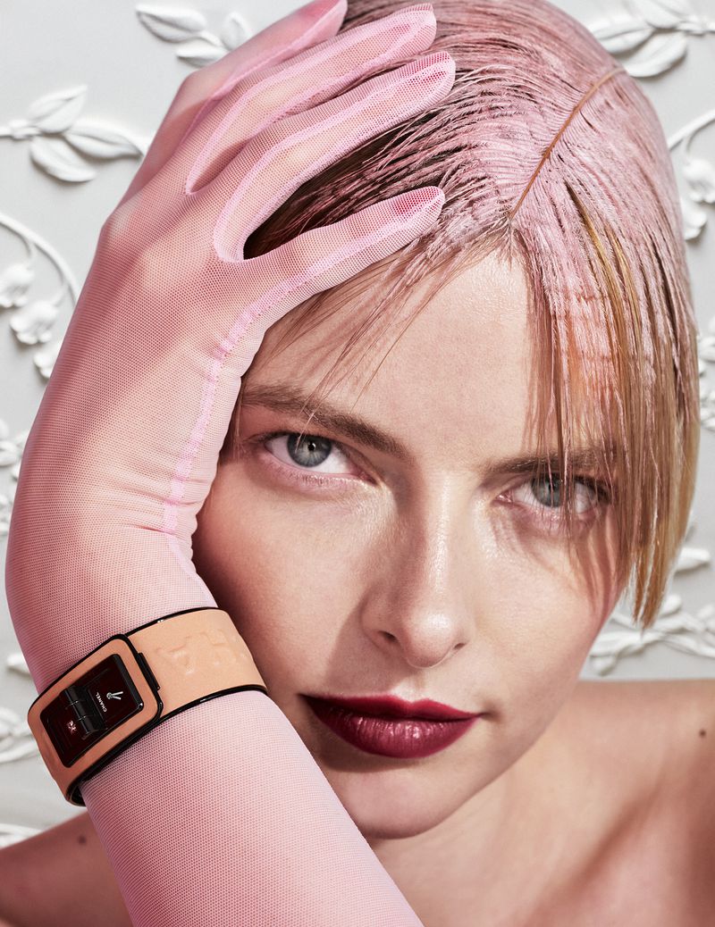 Close-up portrait of a woman with pink tinted hair and light pink glove, wearing a tan and black watch against an ornate white background.