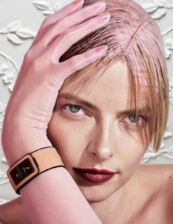 Close-up portrait of a woman with pink tinted hair and light pink glove, wearing a tan and black watch against an ornate white background.
