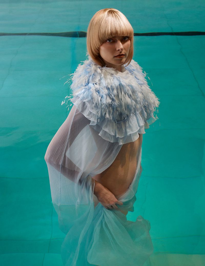 Blonde woman with a feathered collar and sheer fabric submerged in turquoise water, portrait-oriented shot, beauty and fashion style.