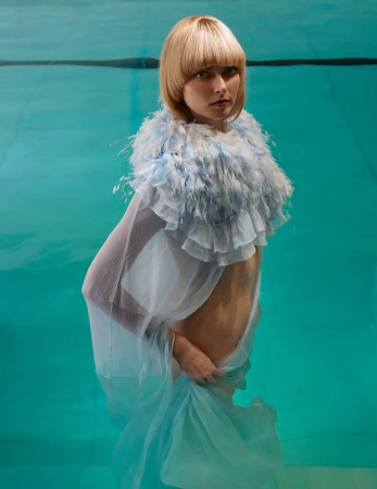 Blonde woman with a feathered collar and sheer fabric submerged in turquoise water, portrait-oriented shot, beauty and fashion style.