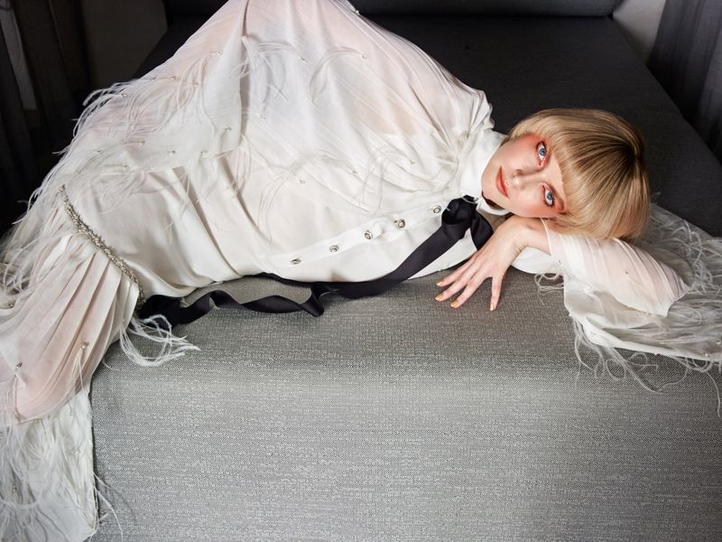 Blonde female model in a white feathered gown lying on a grey sofa, looking at the camera with direct gaze and neutral expression.