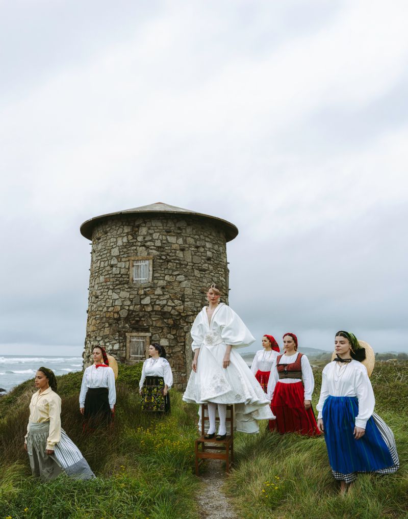 Outdoor fashion shoot features seven women in traditional dresses and headscarves, posing on a grassy hill with a stone windmill.