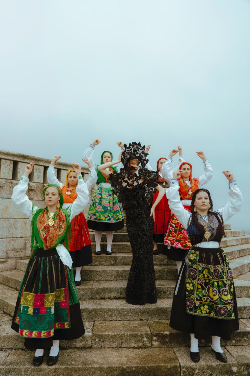 Group of women in traditional dress raising arms in outdoor setting on stone steps, neutral sky, lace details, black and green