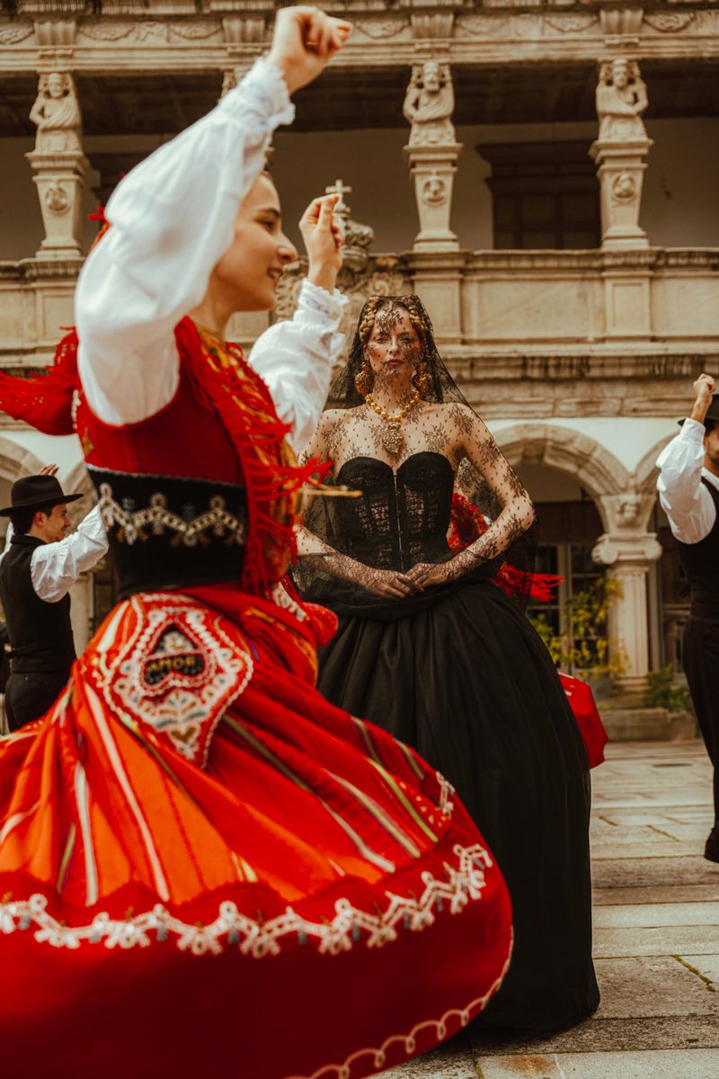 Two women in traditional clothing dancing outdoors in a courtyard, surrounded by a group of men in hats and waistcoats.