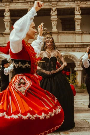 Two women in traditional clothing dancing outdoors in a courtyard, surrounded by a group of men in hats and waistcoats.