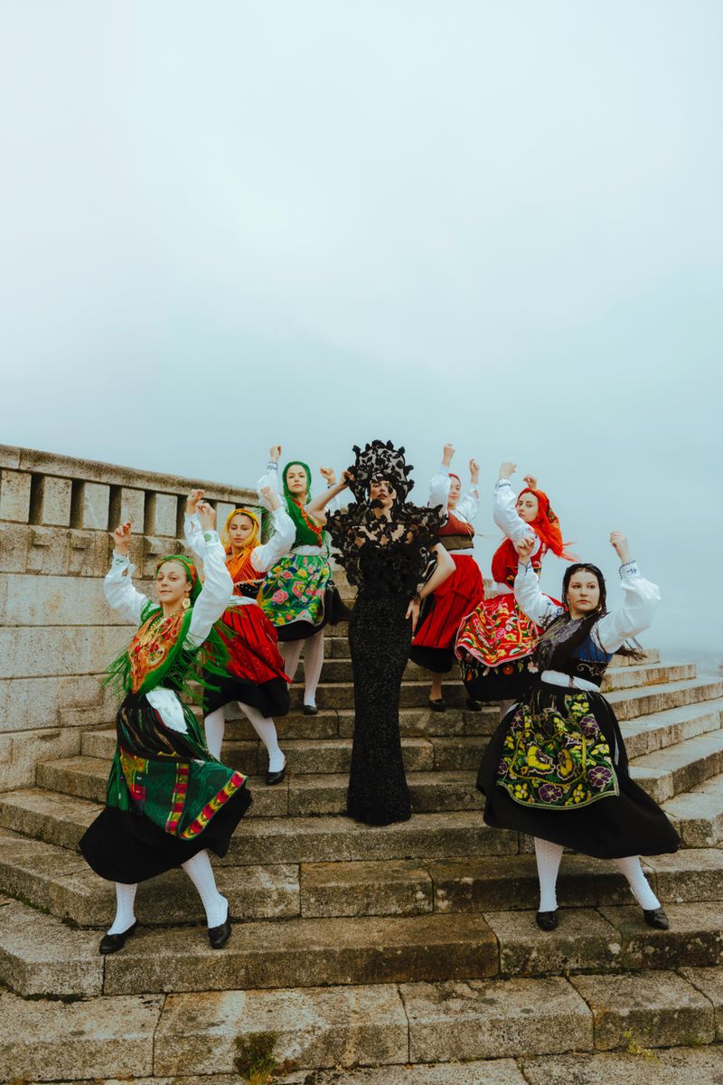 Group of women in traditional folk costumes standing on stone steps with arms raised against an overcast sky in an outdoor setting.