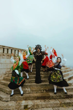 Group of women in traditional folk costumes standing on stone steps with arms raised against an overcast sky in an outdoor setting.