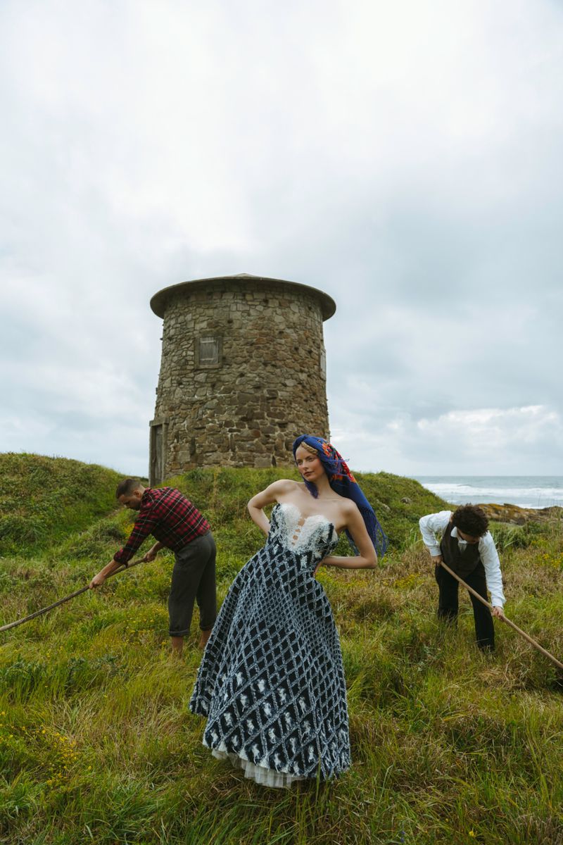 A vintage fashion shot features a model in a patterned dress with two figures with rakes in a grassy field by a stone tower.